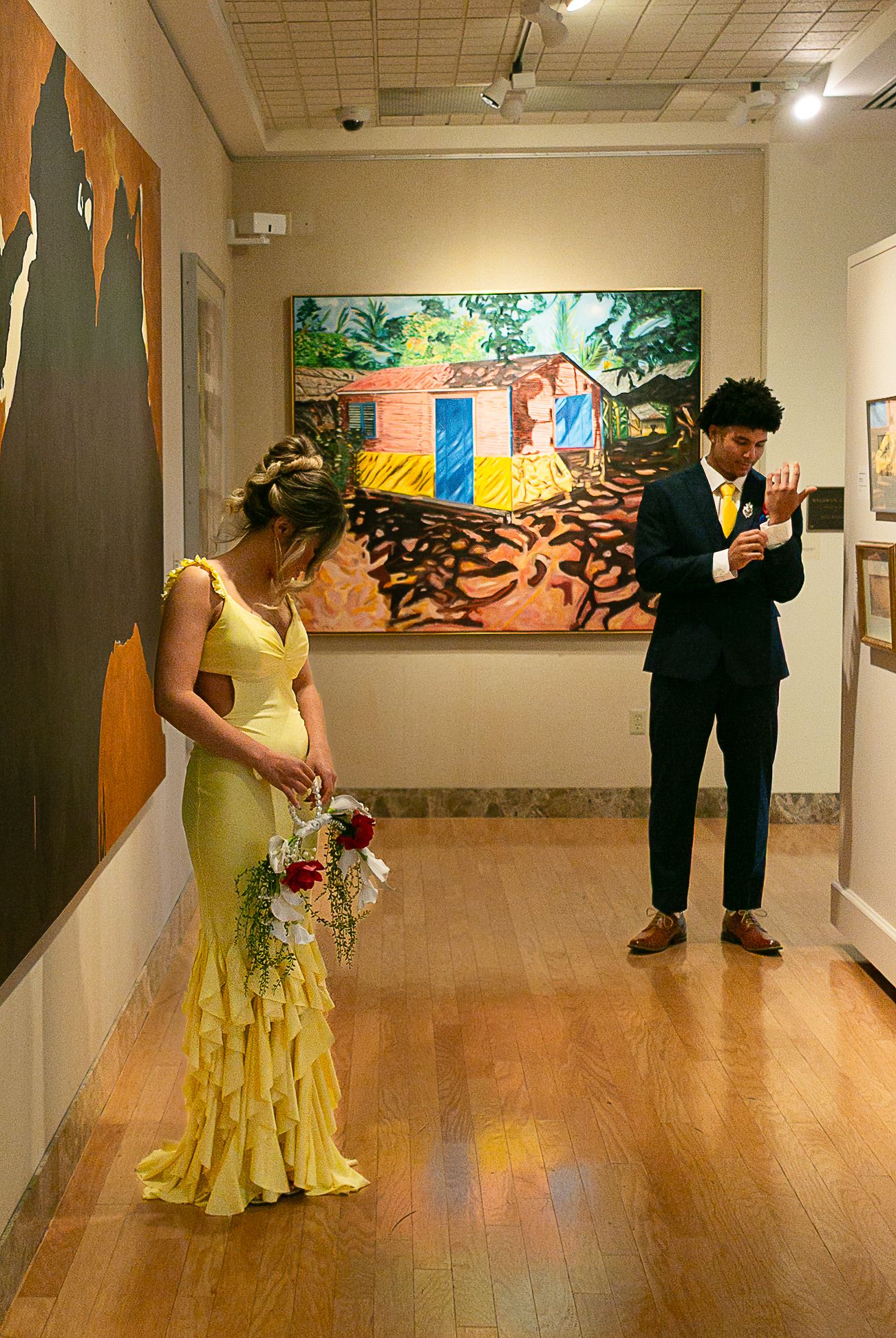 Couple in wedding attire poses in art gallery, bride in yellow ruffled gown holding bouquet, groom in navy suit reading vows.