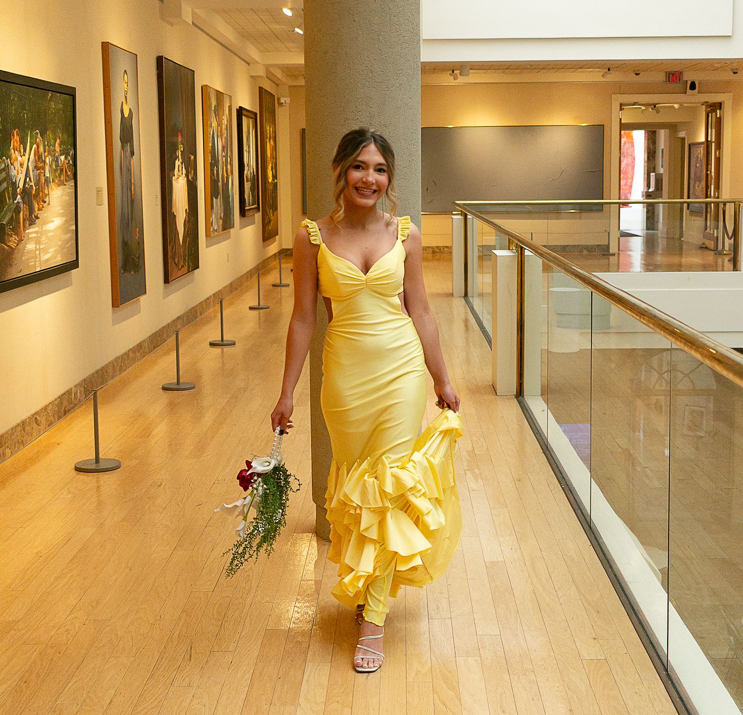 Woman in yellow ruffled mermaid gown holding red bouquet walking through art gallery corridor.