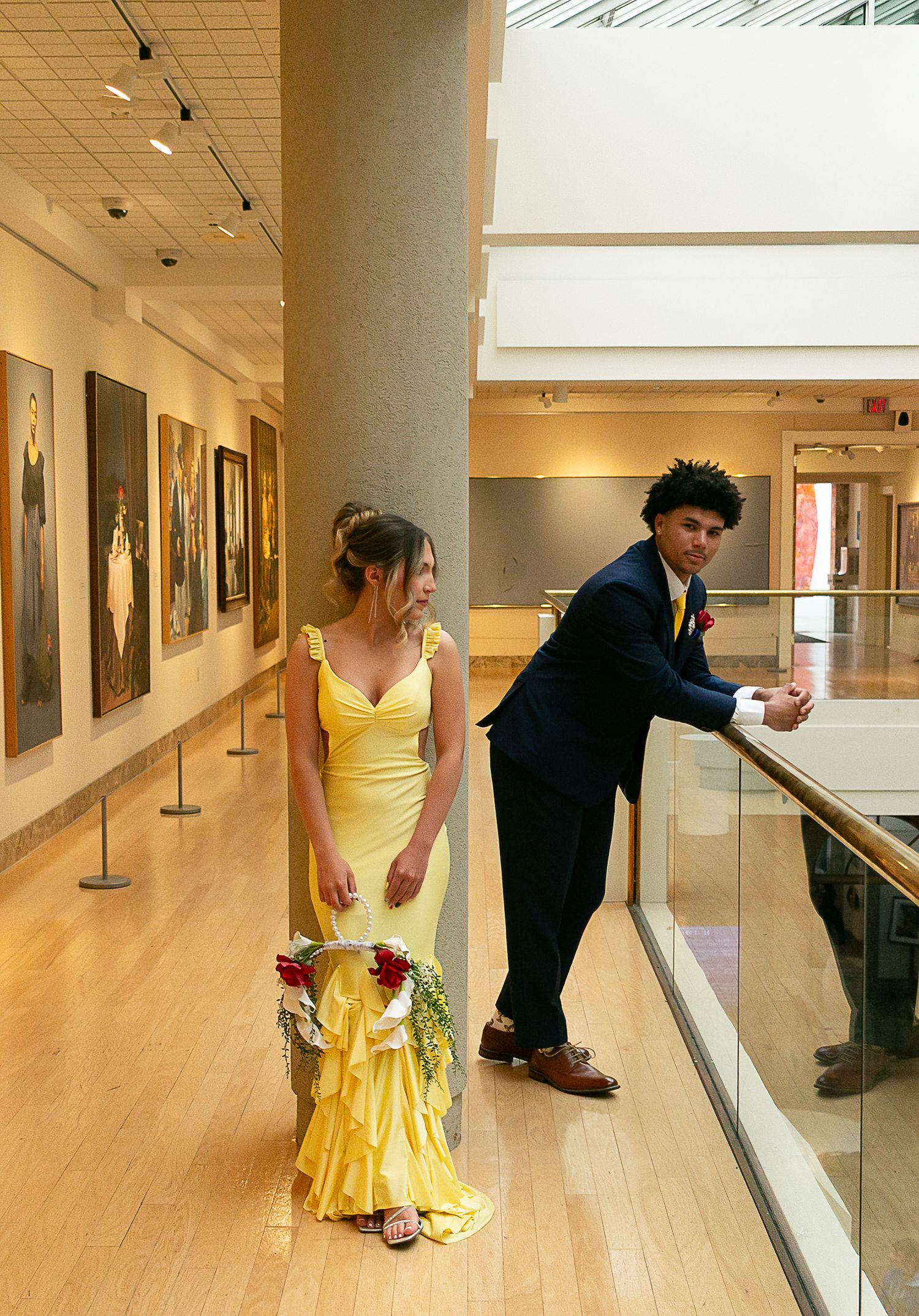 Couple in formal wear poses in art gallery, woman in yellow gown holds bouquet, man in navy suit leans on railing.