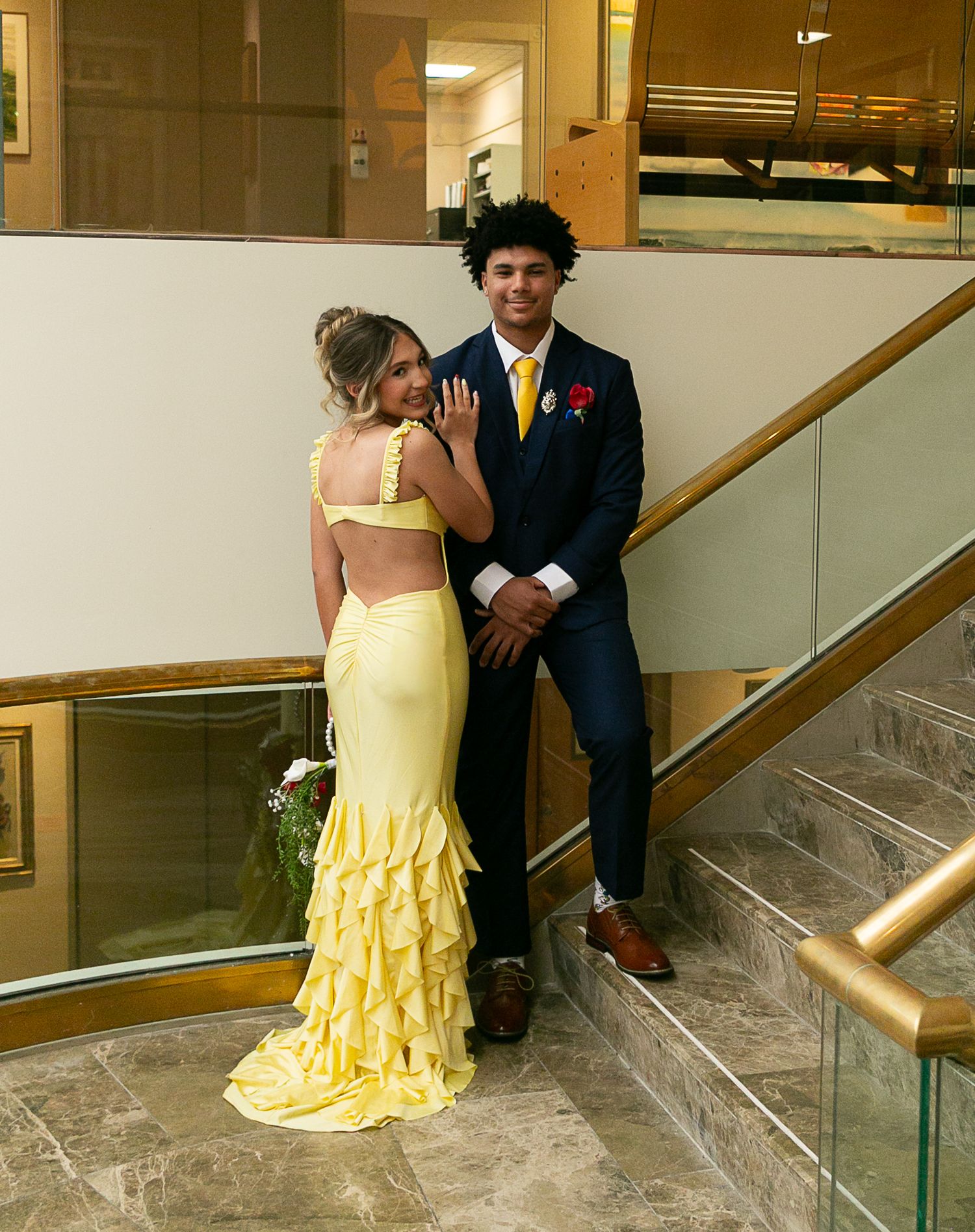 Couple in matching yellow accessories poses on marble stairs, woman in yellow ruffle gown, man in navy suit.
