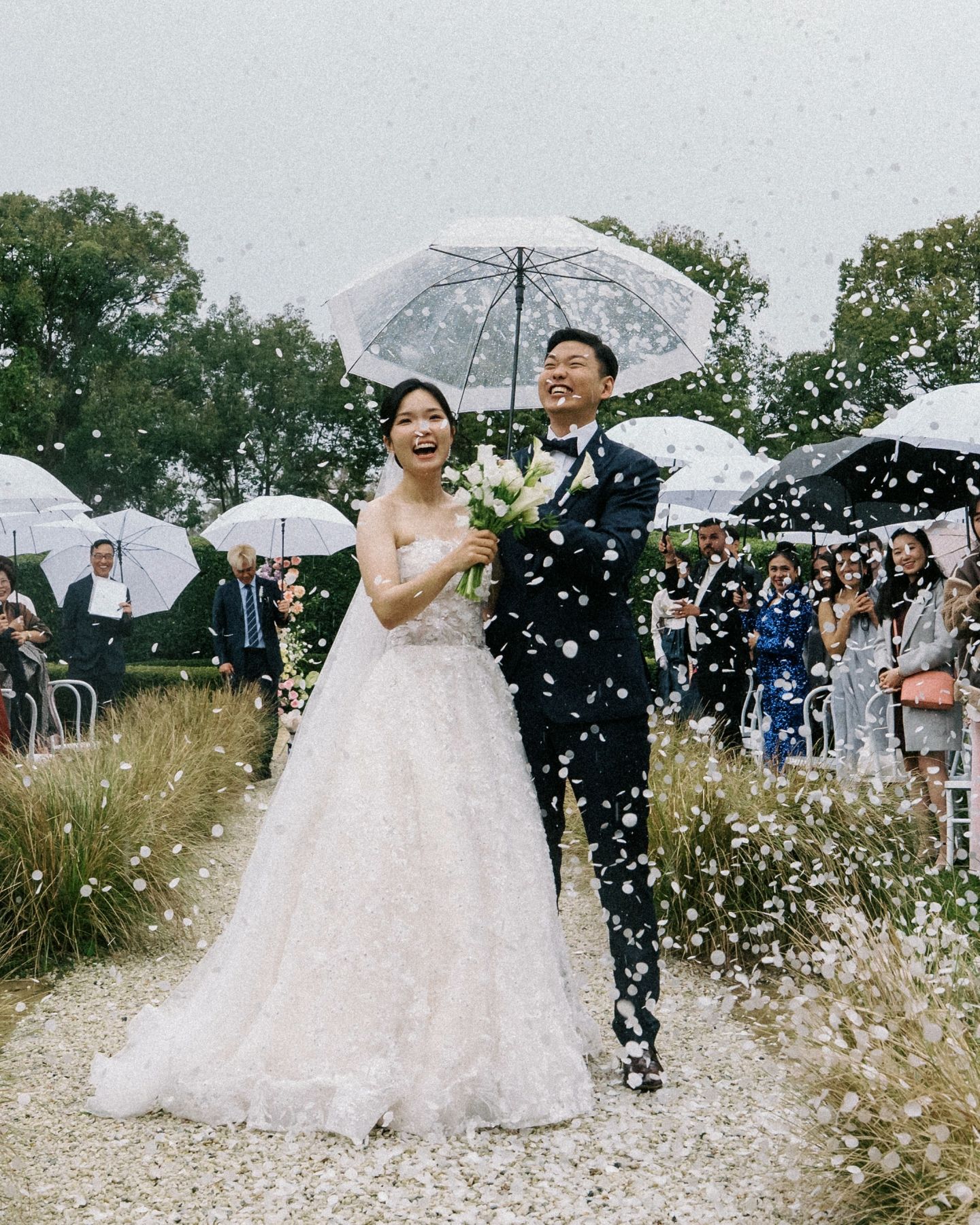 An intimate outdoor wedding ceremony under oak trees with white umbrellas shielding guests from light rain.