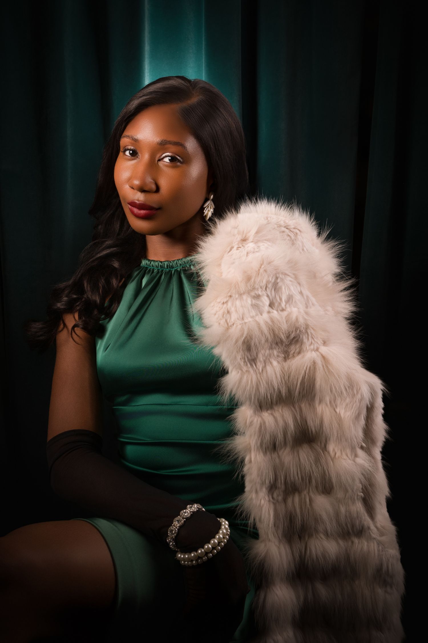 Elegant Black woman in green satin dress holding white fur stole, wearing pearl bracelet, posed against teal curtain backdrop.