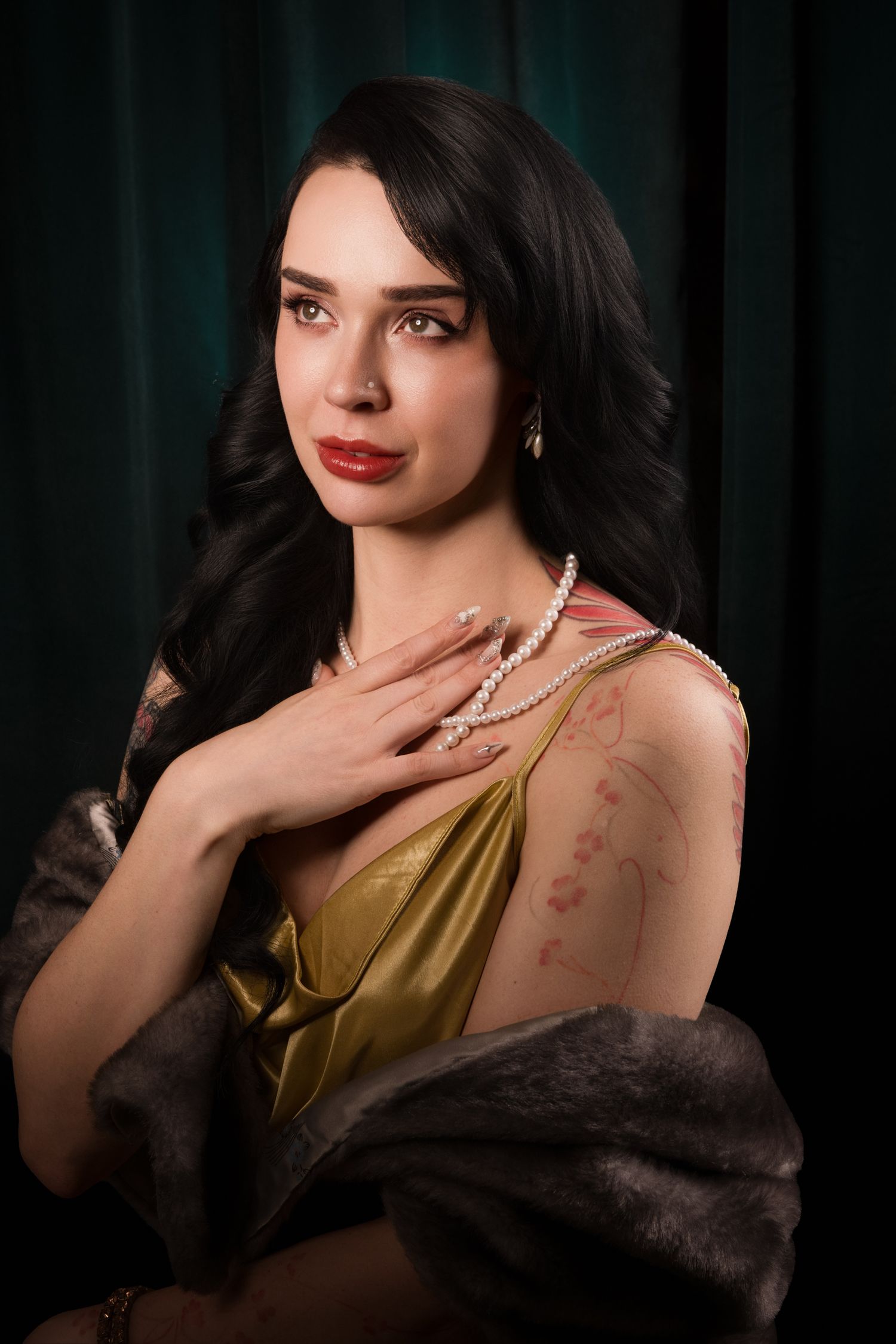 Elegant woman in gold dress with fur stole, red lips, dark hair, pearl earrings, posing against dark green backdrop.