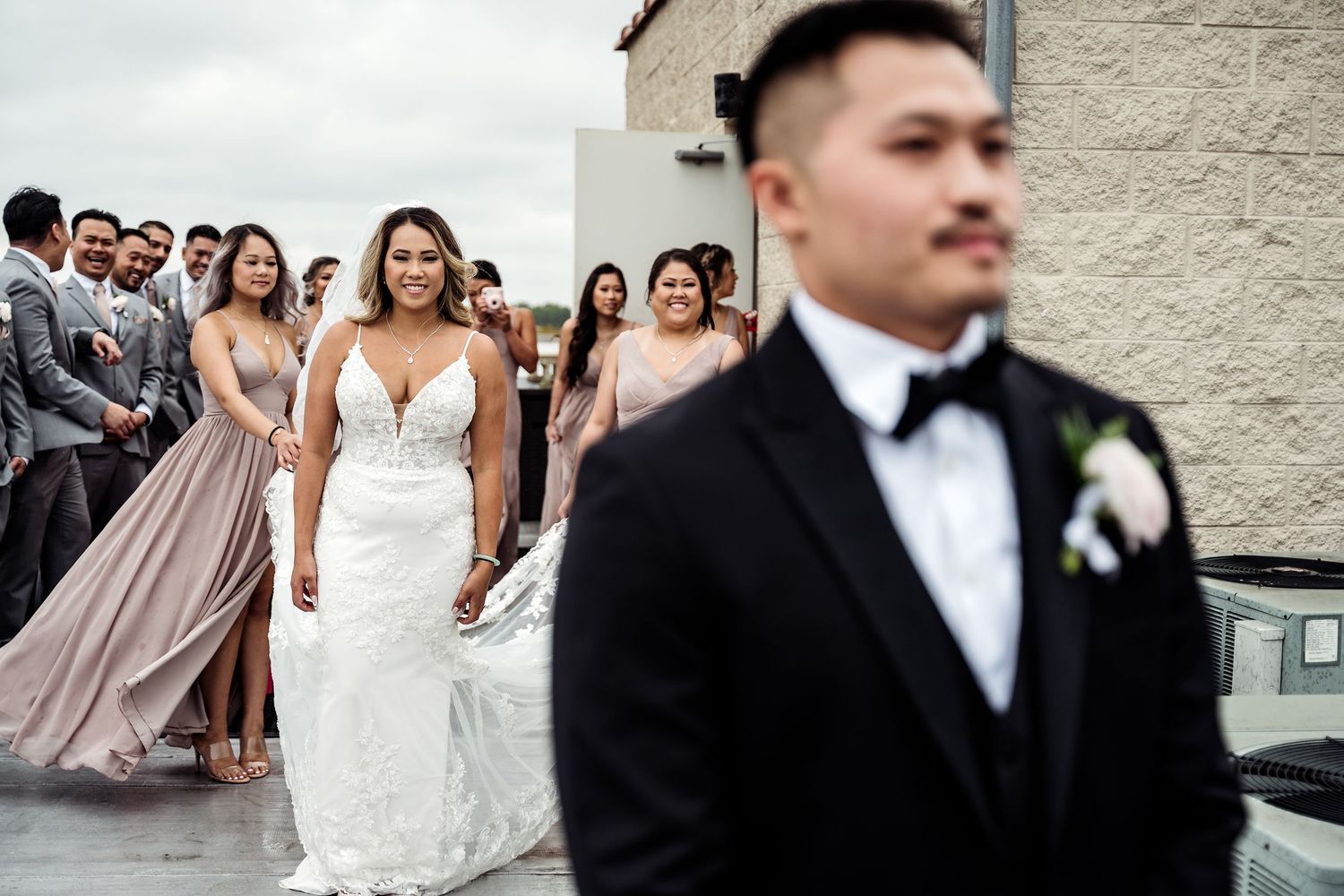 Bride in white lace gown walks toward groom in black tuxedo with bridal party during outdoor first look.