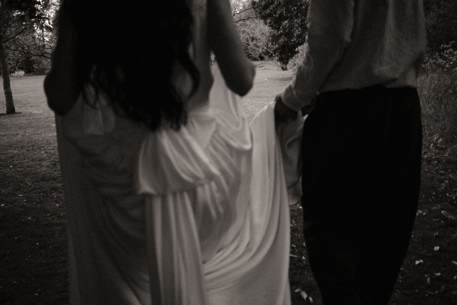 Groom carrying bride in white dress, laughing joyfully outdoors among lush green trees.