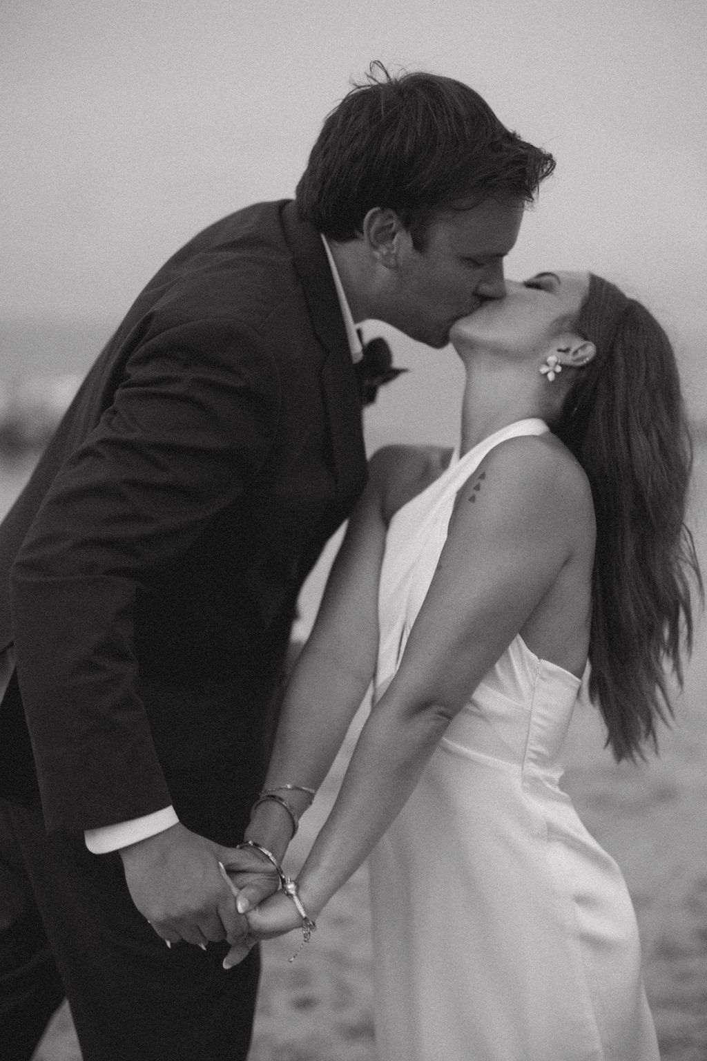 Black and white photo of a bride and groom sharing a romantic kiss on the beach, holding hands.