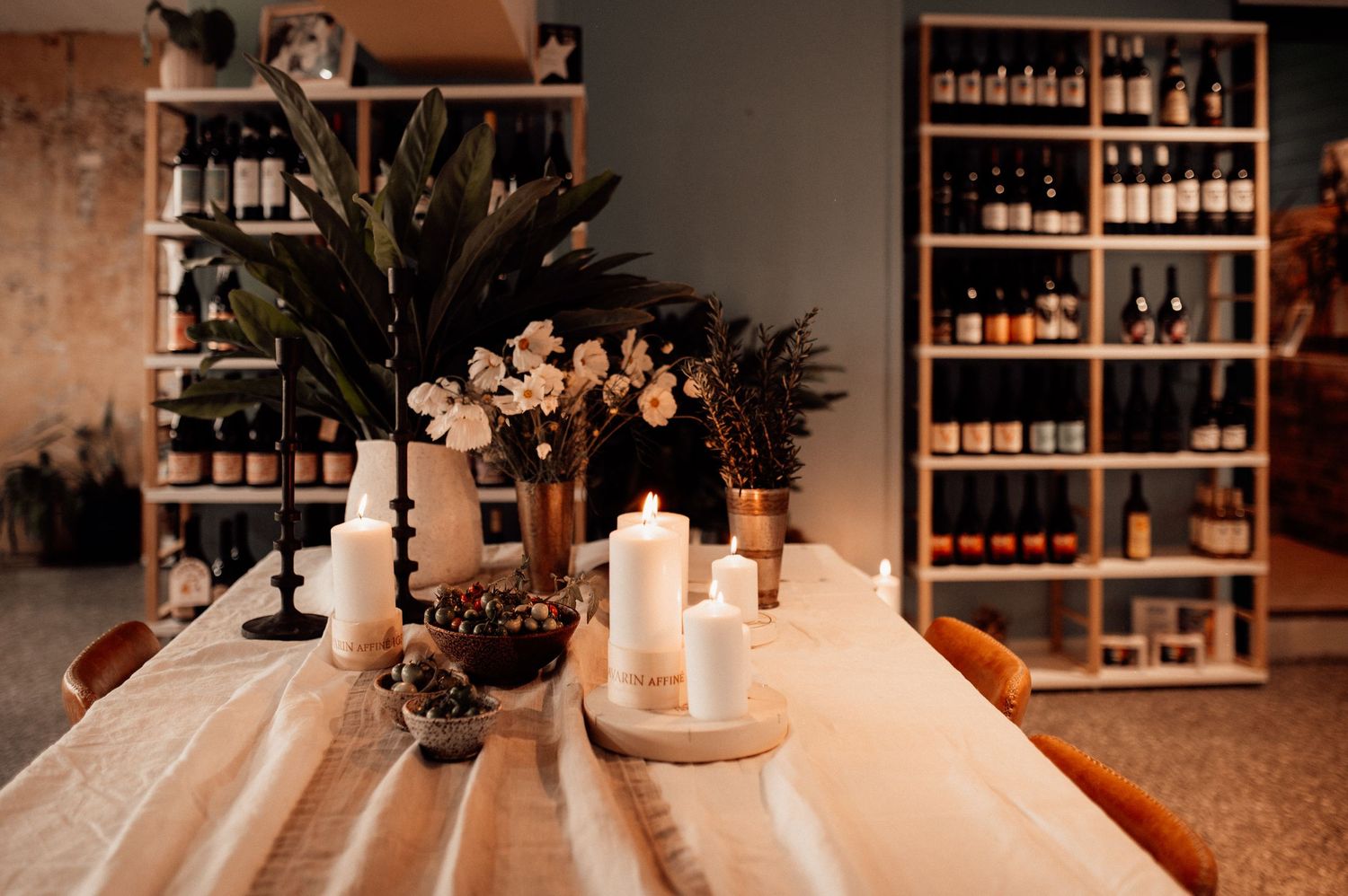 Elegant wooden table with white pillar candles, floral arrangements, and wine bottle shelving in a cozy wine bar setting.