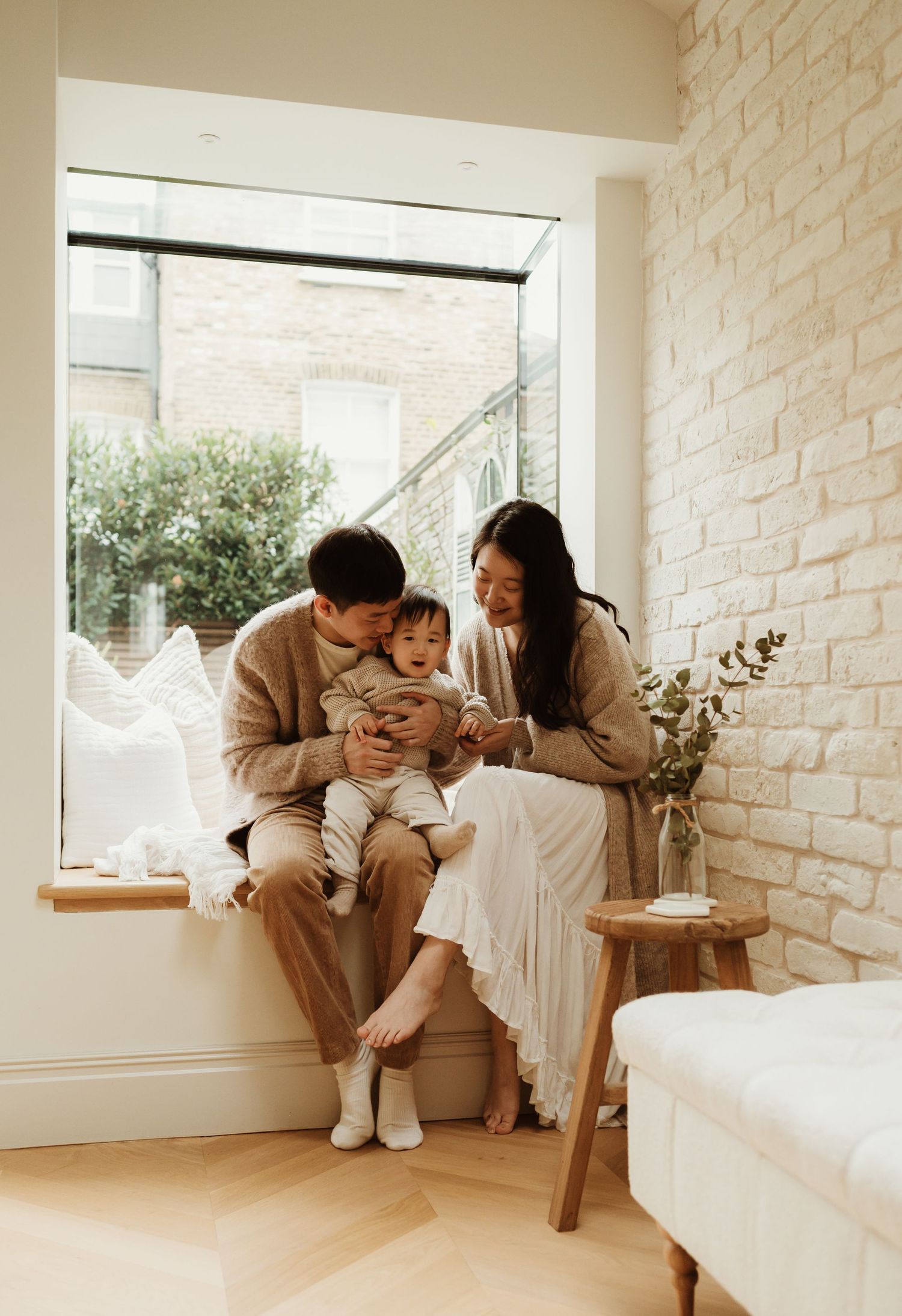 Mother and child sitting on a window seat in a bright, modern home with exposed brick walls and garden views.