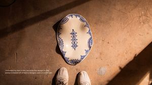 Overhead view of ornate blue and white ceramic plate with Henna-inspired designs on rustic floor beside white shoes.