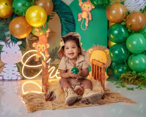 Toddler in safari outfit sits before jungle-themed birthday backdrop with animal cutouts.