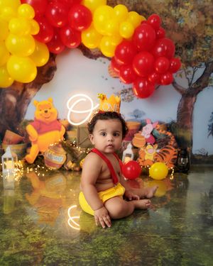 Baby in yellow outfit sits before Winnie the Pooh birthday backdrop with red and yellow balloons.