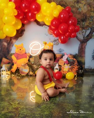 Baby in yellow suspenders sits before Winnie the Pooh backdrop with red and yellow balloon arch.