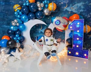 Toddler in astronaut costume poses with rocket and number 1 prop under space-themed balloon arch.