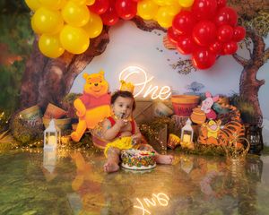Baby girl in yellow dress smashes Winnie the Pooh birthday cake under red and yellow balloon arch.