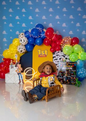 Baby dressed as Woody sits in wooden bed with Toy Story themed colorful balloon arch and character props.