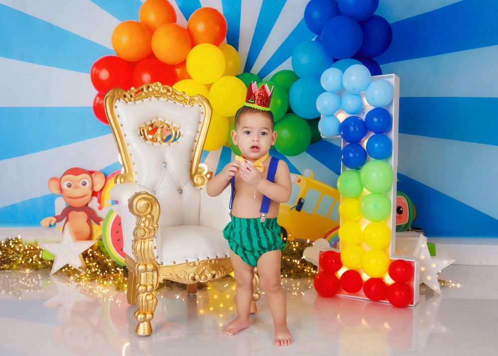 A baby boy in a watermelon outfit stands before a white and gold throne chair with a vibrant rainbow balloon display.