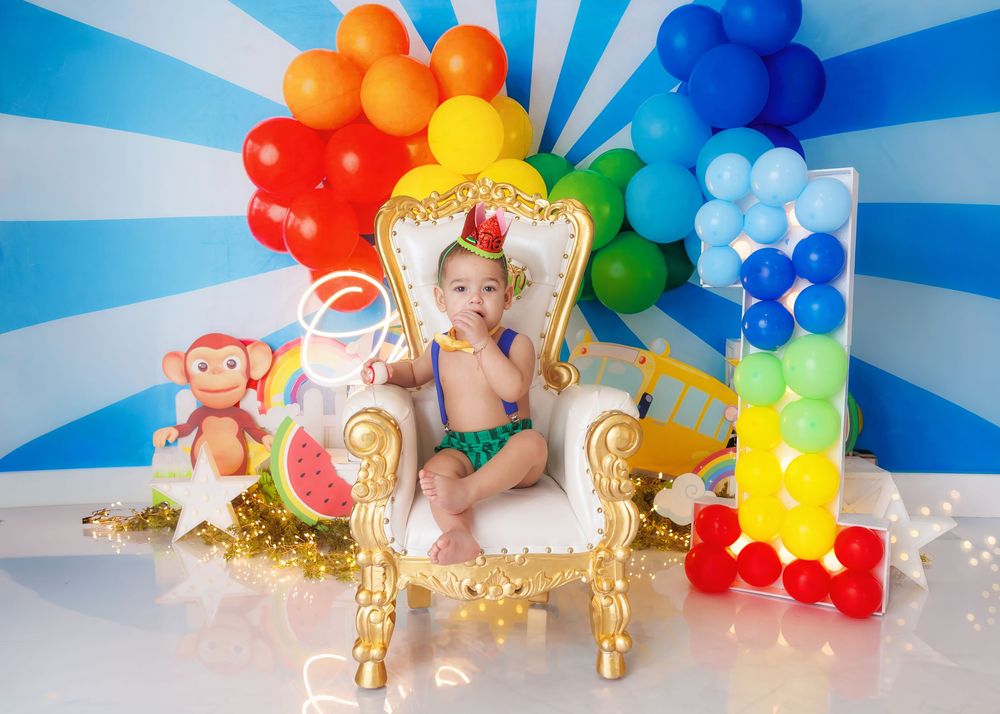 A baby boy in a colorful outfit sits on a gold and white throne chair with a rainbow balloon arch and number one decor.