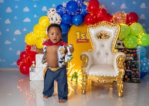 Baby boy dressed as Woody from Toy Story stands by golden throne at colorful first birthday party.