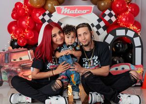 A family sits together in front of a Mateo Disney Cars themed birthday party backdrop with red and gold balloons.
