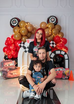 A family of three poses in front of a Disney Cars themed birthday backdrop with red and gold balloons.