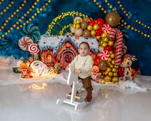 Toddler stands on white rocking horse before a festive gingerbread house backdrop with candy canes, colorful balloons and holiday decor.