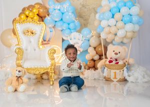 Smiling baby boy sits before blue and gold balloon arch with teddy bears and throne chair at 1st birthday.