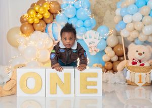 A baby boy stands behind large ONE letter blocks with a blue and gold balloon garland and teddy bear decorations behind him.