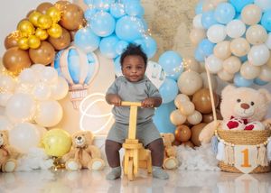 Baby boy on wooden bike toy surrounded by blue, gold, and white balloon garland with teddy bears for 1st birthday.