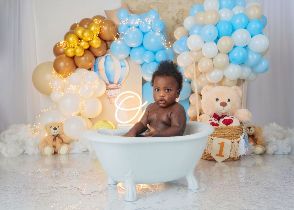 A baby boy sits in a white bathtub surrounded by blue and gold balloons, teddy bears, and a hot air balloon decoration.