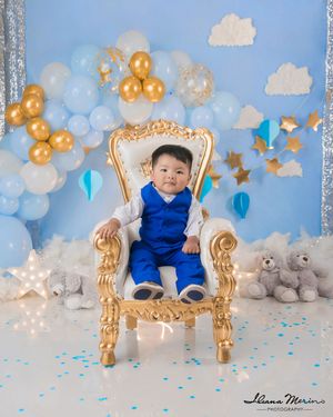 Baby boy in blue royal outfit sitting on gold throne with blue balloons and teddy bears.