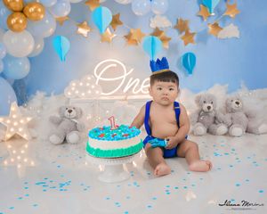 Baby boy with blue crown celebrating first birthday with cake surrounded by teddy bears and balloons.