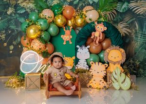 Baby in tan outfit sits on wooden bed surrounded by jungle animal cutouts at a safari birthday party.