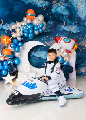 Young boy in astronaut costume sits on inflatable space shuttle at a space-themed birthday celebration.