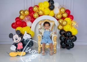 Baby in denim overalls sits on a gold throne chair surrounded by Mickey Mouse themed red, yellow and black balloon arch.