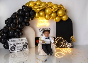 A baby boy sits in a chair at a Carter hip-hop themed first birthday party with black and gold balloon arch.