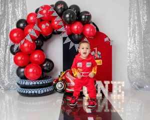 Toddler in racing suit celebrates birthday with red and black balloon arch backdrop.