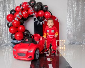 Child in red racing suit stands beside toy car at race car birthday party.