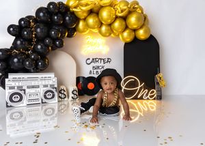 A baby boy poses at a Carter hip-hop music themed first birthday party with black and gold balloon arch decor.