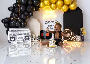 A baby boy sits with a smash cake at a Carter hip-hop music themed first birthday party with black and gold decor.