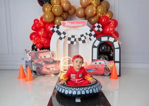 A baby boy in a red outfit sits in a tire at a Mateo Disney Cars themed first birthday cake smash setup.