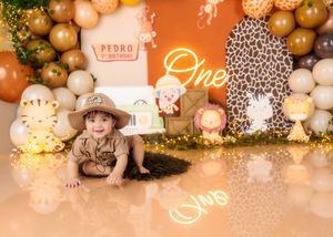 Baby in safari outfit crawls on floor at Pedro's jungle-themed first birthday party with neon One sign.