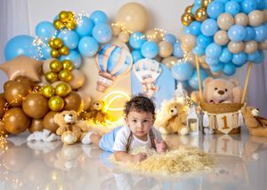 Baby lies on floor surrounded by teddy bear themed blue, gold balloon arch with hot air balloon decoration and stuffed animals.