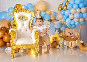 Baby stands by ornate gold throne with blue and gold teddy bear themed balloon arch and hot air balloon decoration with number one.