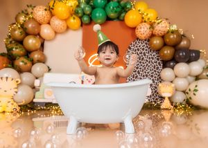 Smiling baby in a white bathtub surrounded by jungle-themed balloons and animal toys at a birthday party.