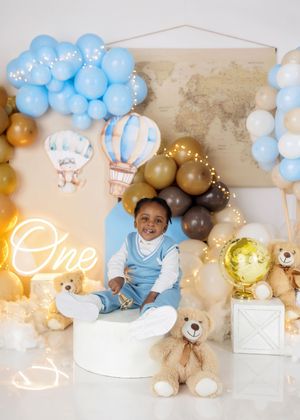 Baby boy in blue outfit sits among teddy bears and balloons at a travel-themed first birthday celebration.