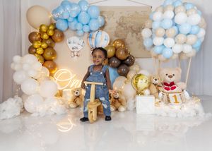 Toddler in denim overalls stands beside wooden bike with blue, gold, and white balloon arch and teddy bears.