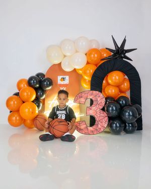 Boy in basketball uniform sits with ball before orange black balloon arch and number 3 prop.