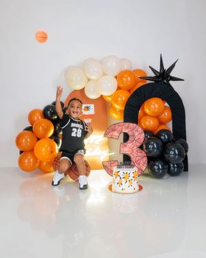 Boy in basketball jersey jumps beside orange and black balloon arch with number 3 birthday prop.