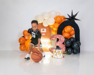 Boy in basketball uniform dribbles ball beside orange black balloon arch and number 3 prop.