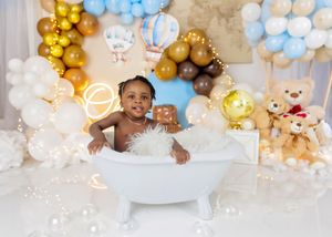 Baby sits in white bathtub surrounded by blue, gold, and brown balloon arch with teddy bear decorations.